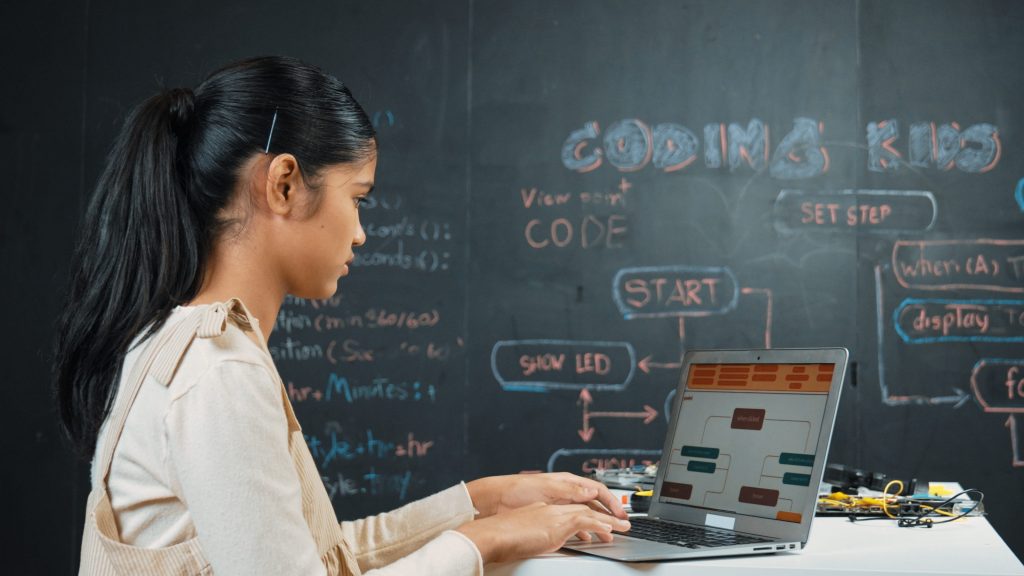 Female student coding on a laptop with a flowchart on the chalkboard behind her