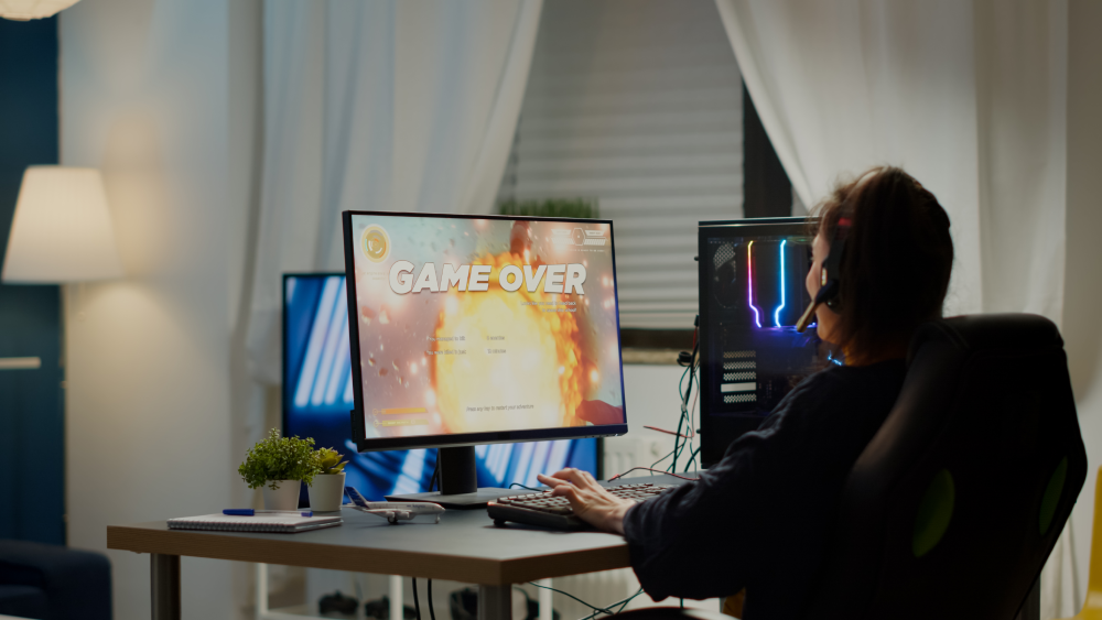 A game developer at an RGB gaming PC setup with a game over screen displayed on the monitor