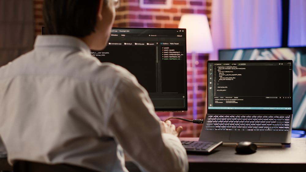A software developer writing C++ game code across dual monitors in a dimly lit home office