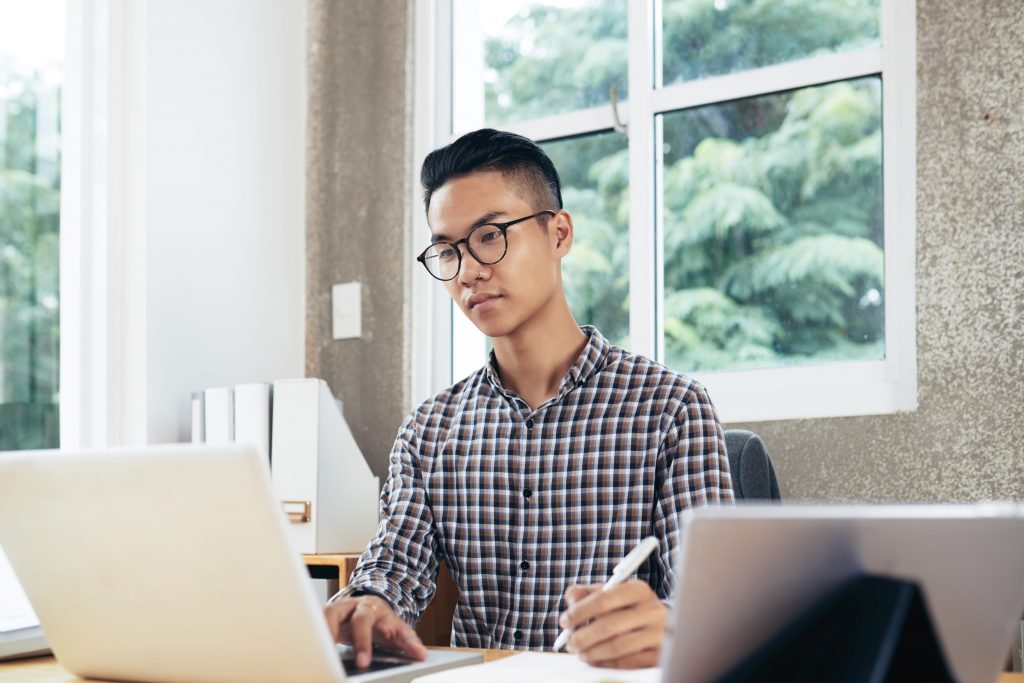 Student taking notes while working on a laptop in a bright home