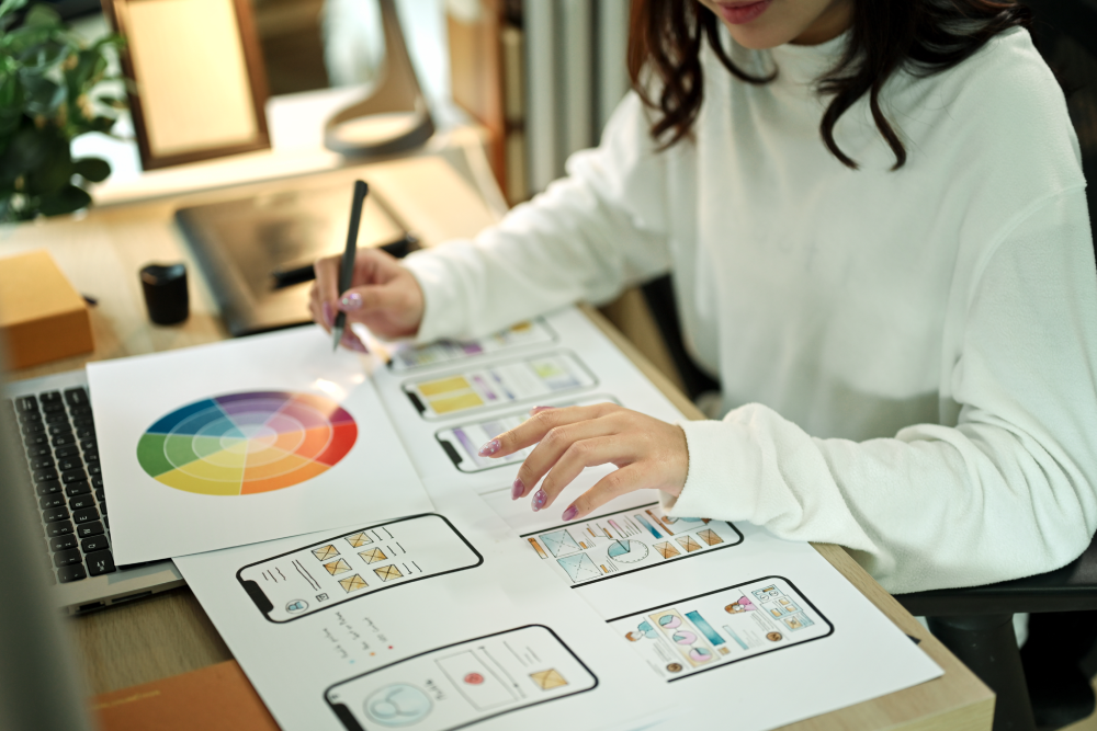 A UX designer reviewing printed mobile app wireframes and a color wheel at her desk